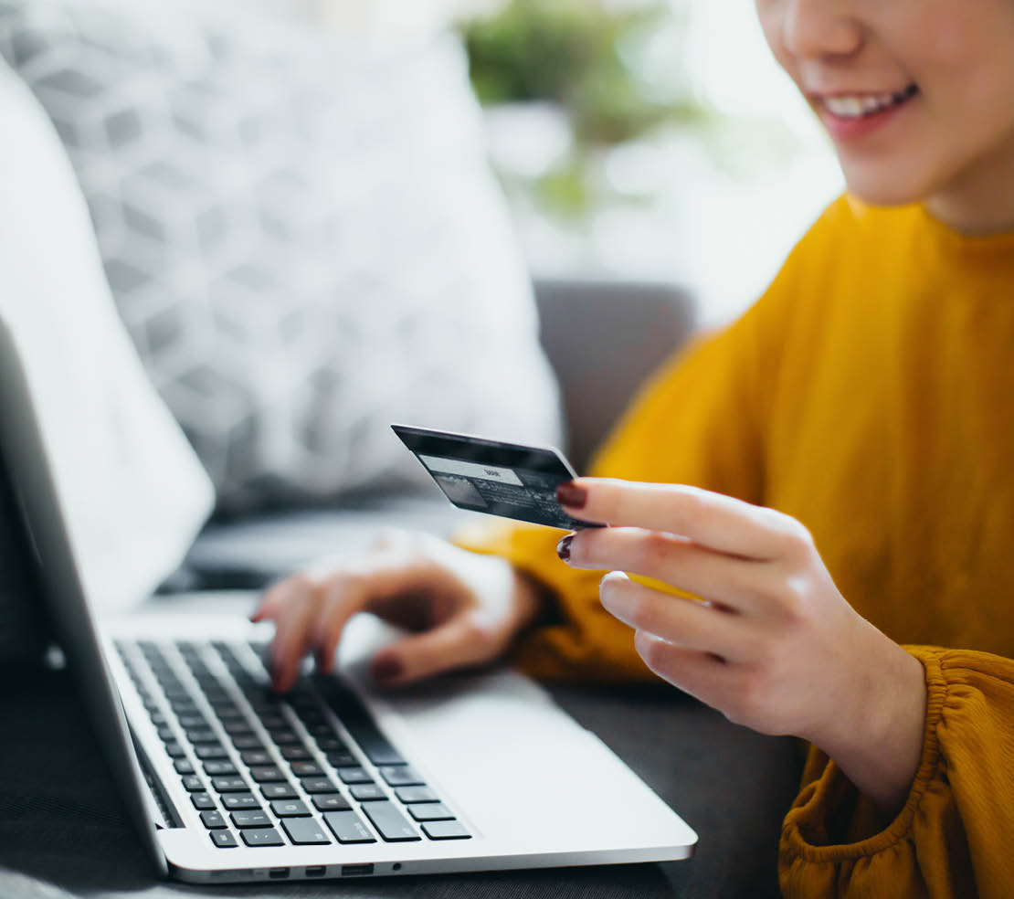 Close up of beautiful smiling young Asian woman sitting on the floor by the sofa, managing financial bill payment with laptop and credit card on hand at cozy home. Technology makes life so much easier