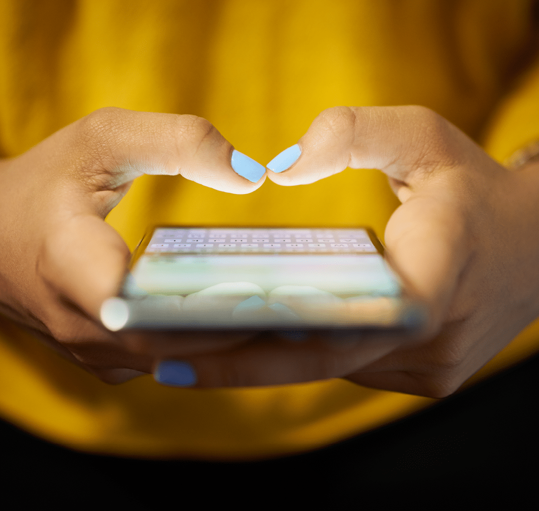 Young woman using cell phone to send text message on social network at night. Closeup of hands with computer laptop in background